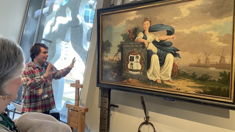 Een man legt een schilderij uit dat een vrouw, een haan en molens langs een kust toont.