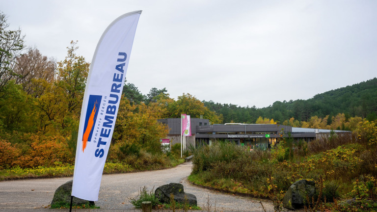 Een stembureauvlag van de gemeente Bergen bij een gebouw omgeven door herfstbomen en een bosrijke omgeving.