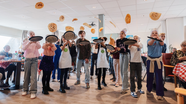 Kinderen bakken en gooien pannenkoeken de lucht in met koekenpannen in een zaal met toekijkende volwassenen.