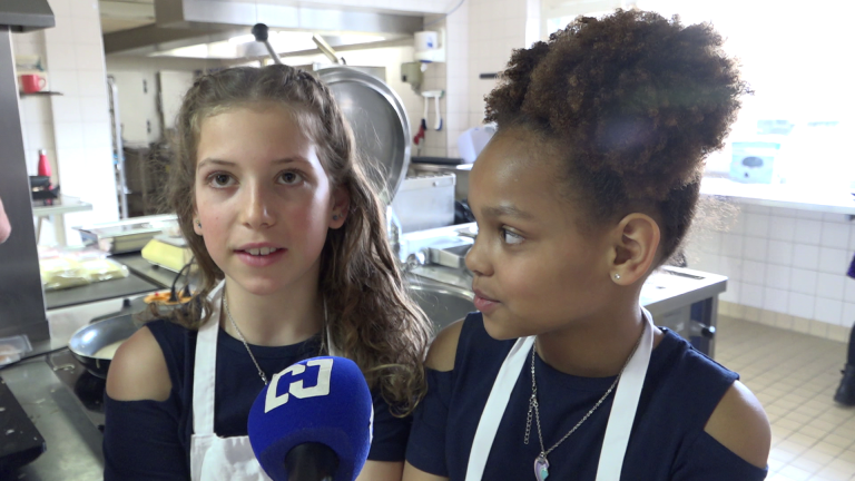 Twee meisjes in een keuken, dragen schorten en staan voor een microfoon met een blauw logo.