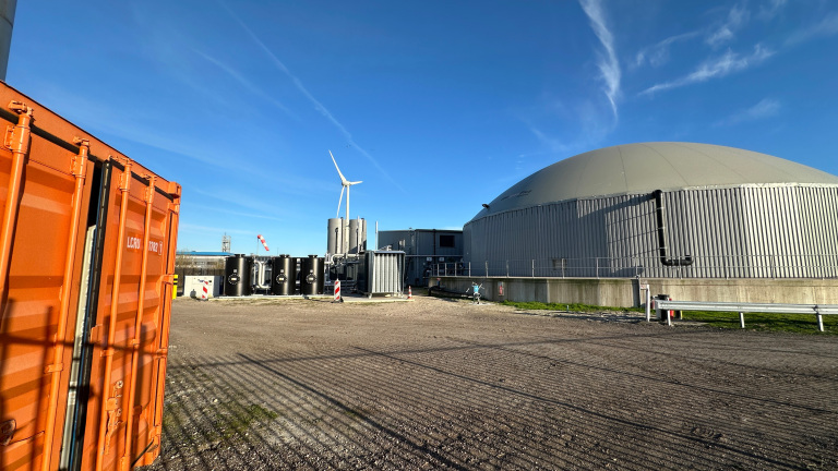 Industrieel terrein met een oranje container, windmolen, grote ronde loods en pijpleidingen onder een blauwe lucht.