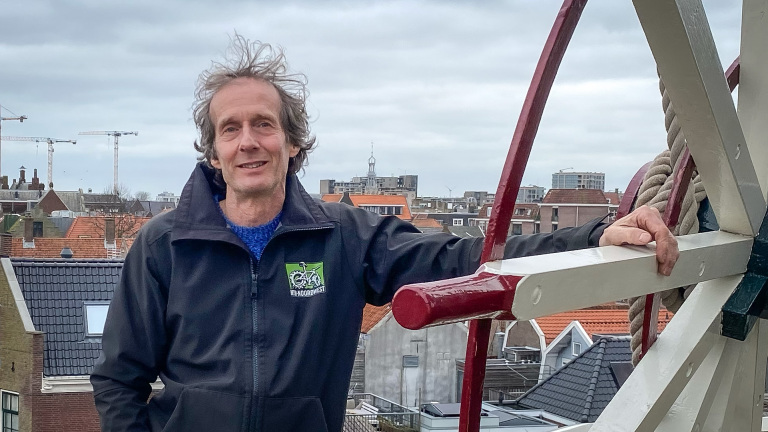 Man in zwarte jas met groen logo staat buiten naast een molenrad, met een stad op de achtergrond.
