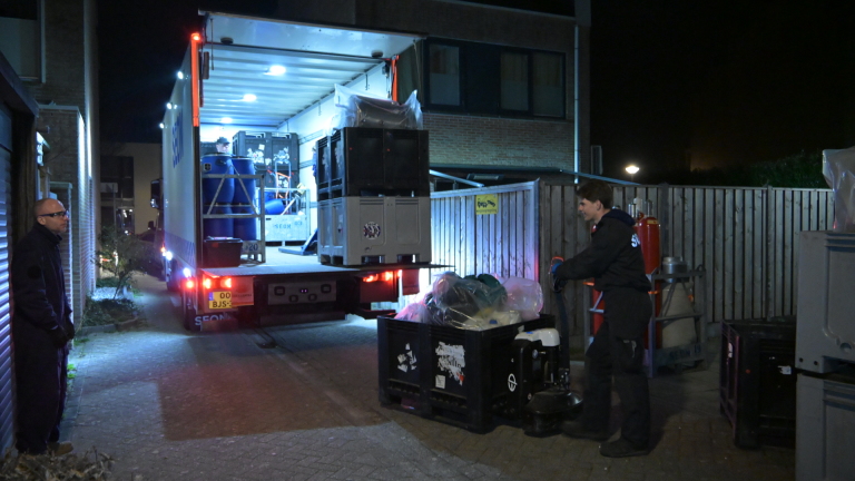 Twee mannen laden 's nachts een vrachtwagen uit met behulp van kratten en vaten in een smalle straat.