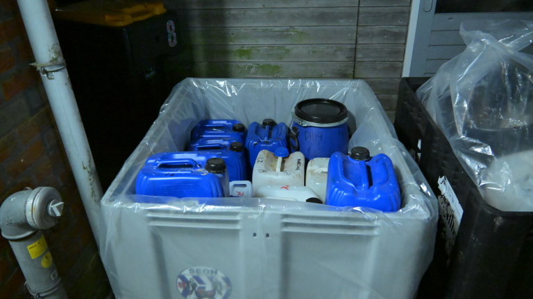 Plastic container met blauwe en witte jerrycans en vaten, opgeslagen in een met folie beklede bak in een donkere ruimte.