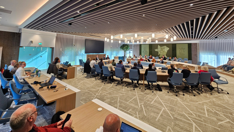 Mensen zitten in een moderne vergaderzaal met blauwe stoelen en houten tafels, diverse laptops op tafel.