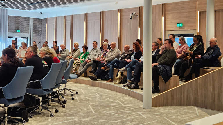 Mensen zitten in een moderne vergaderruimte met gestoffeerde stoelen en een houten afwerking, aandachtig naar een spreker luisterend.