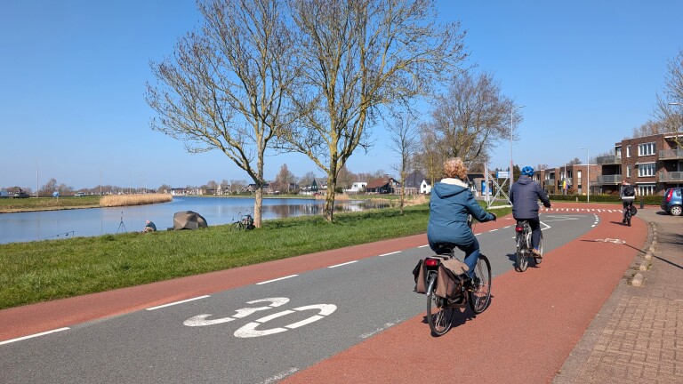 Mensen fietsen langs een kanaal met een visser aan de oever, op een zonnige dag met blauwe lucht.