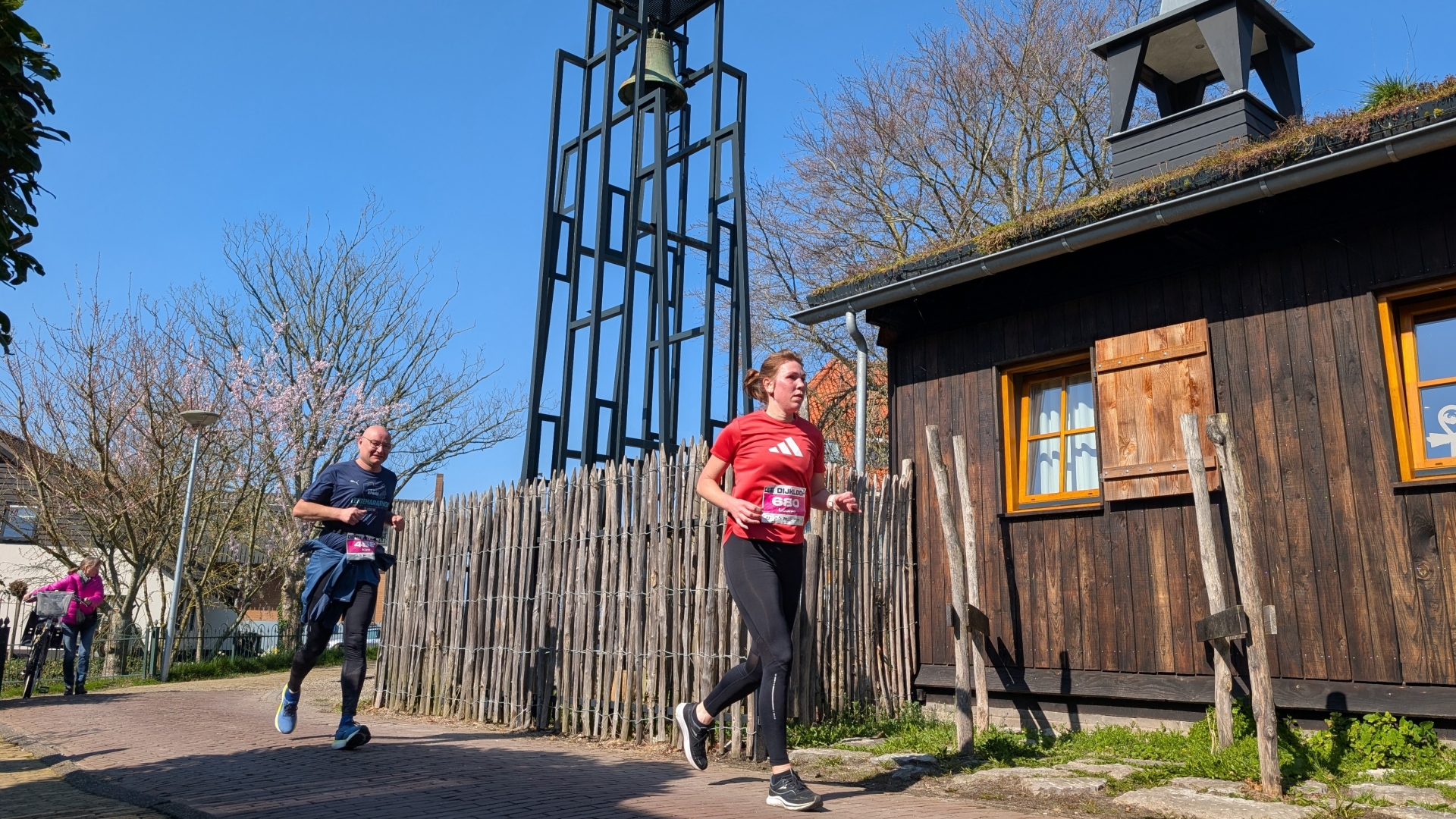 Twee hardlopers rennen langs een houten gebouw en een kloktoren onder een heldere blauwe hemel.