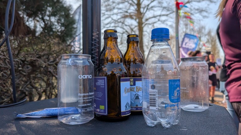 Lege bierflessen en plastic flessen op een tafel, met mensen op de achtergrond in een buitenomgeving.