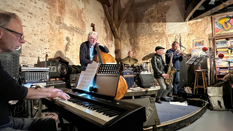 Een jazzband speelt muziek in een rustieke ruimte. Een pianist, contrabassist, drummer, saxofonist en trompettist zijn bezig.