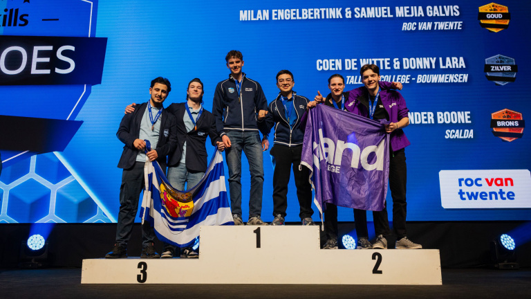 Zes jonge mannen staan op een podium met medailles, vlaggen en een scherm met winnaarsnamen op de achtergrond.