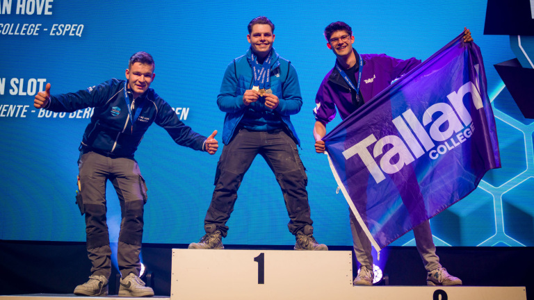 Drie lachende mannen op een podium met een blauwe achtergrond, waarvan één een vlag van Tallant College vasthoudt.