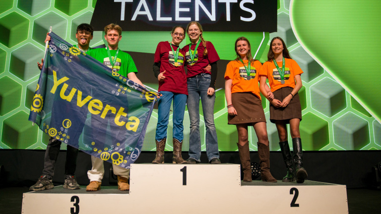 Zes jongeren poseren op een podium; twee dragen een Yuverta-vlag, en anderen dragen medailles en kleurrijke shirts.