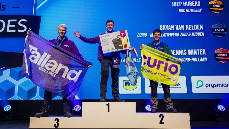 Drie jonge mannen staan op een podium met vlaggen van hun scholen, een van hen houdt een bord vast voor de eerste plaats.