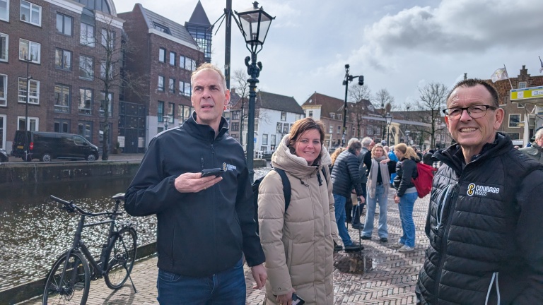 Drie mensen staan buiten op een geplaveide straat nabij een gracht, met fietsen en een groep mensen op de achtergrond.
