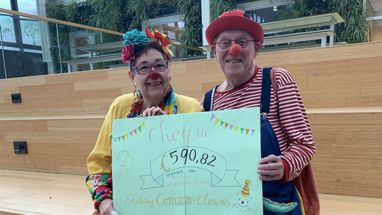 Twee clowns met rode neuzen houden een cheque van €590,82 vast van Stichting Corazón-clowns in een gebouw.