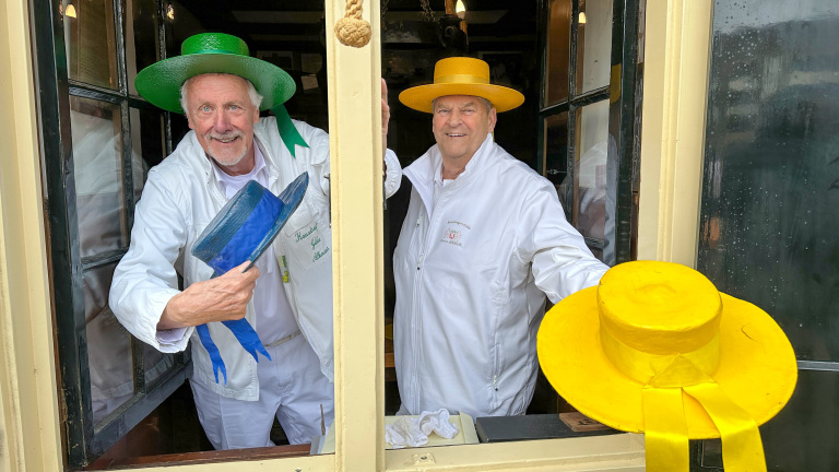 Twee mannen in witte jassen met kleurrijke hoeden, één groen en één geel, staan voor een raam en lachen.