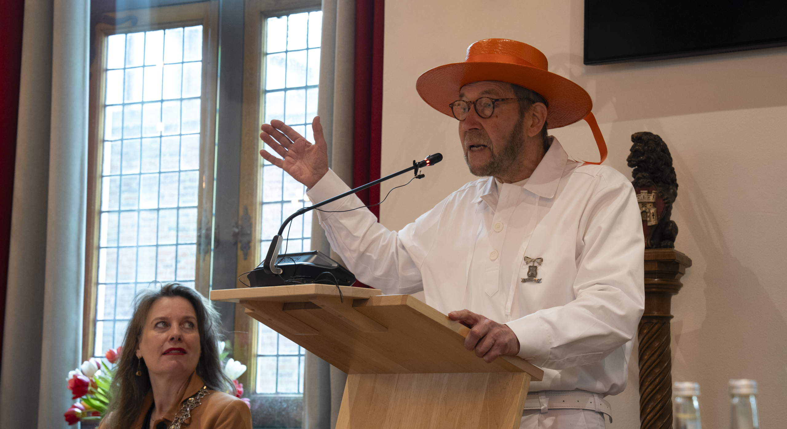 Een man in een witte outfit en oranje hoed houdt een toespraak achter een lessenaar, met een vrouw die aandachtig luistert.
