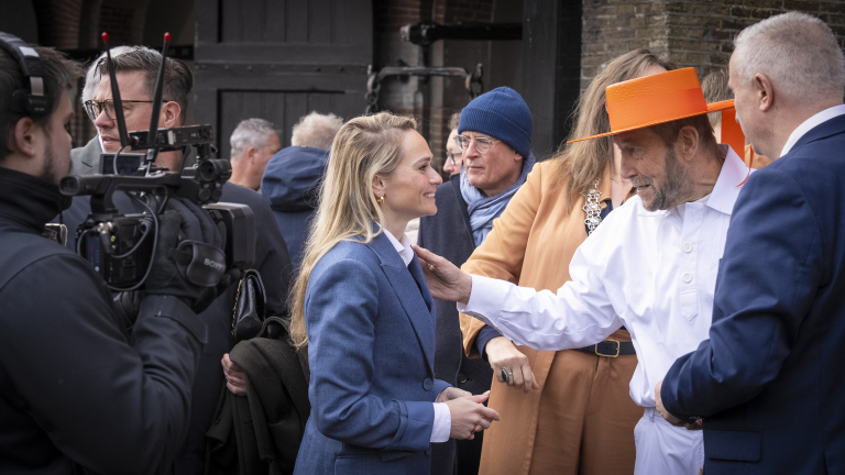 Een groep mensen in gesprek, met een man in een wit pak en oranje hoed die met een vrouw praat, gefilmd door een cameraman.
