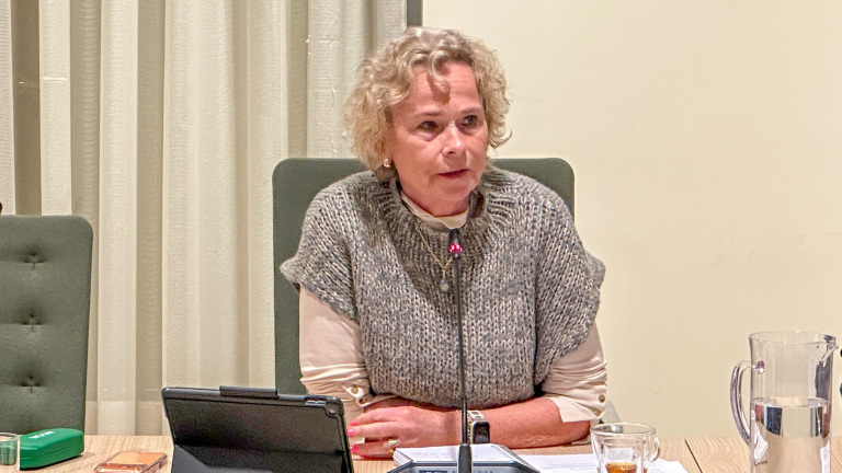 Vrouw met krullend haar zit aan een tafel met microfoon, tablet en documenten voor zich.