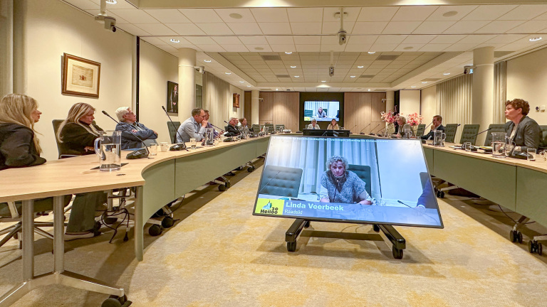 Mensen zitten rondom een vergadertafel met een groot scherm waarop een videoconferentie met een naamplaat zichtbaar is.