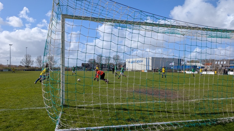 Voetbalwedstrijd met een keeper in rood die een bal probeert te stoppen vanuit het perspectief van het doel, met spelers in geel en groen.