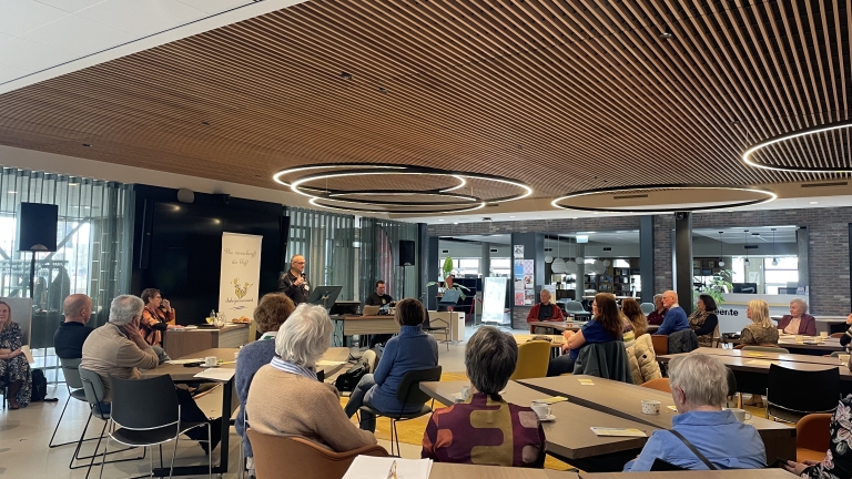 Een groep mensen zit in een moderne zaal en luistert naar een spreker bij een lessenaar.