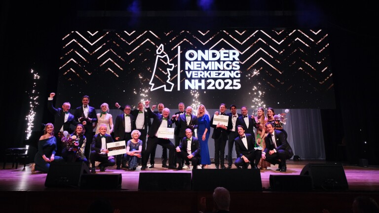 Groep mensen in avondkleding viert een prijsuitreiking op een podium met een groot scherm dat "Ondernemingsverkiezing NH 2025" toont.