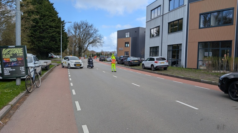 Een verkeersregelaar in een gele outfit staat op de weg en helpt een scootmobiel oversteken, met auto's op de achtergrond.