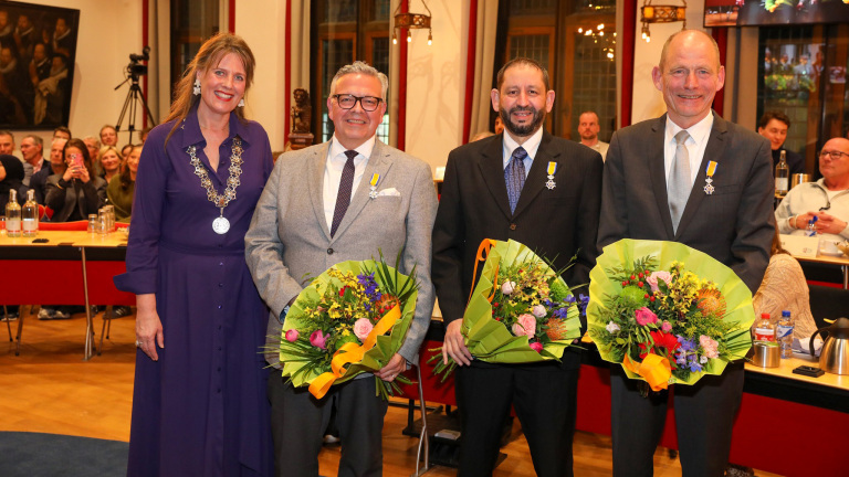 Vier mensen poseren voor een foto in een zaal, waarvan drie mannen bloemen en onderscheidingen dragen.