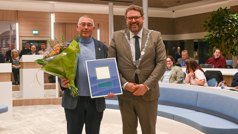 Twee mannen poseren in een zaal; één houdt bloemen en een ingelijste oorkonde vast, terwijl de ander een ambtsketting draagt.