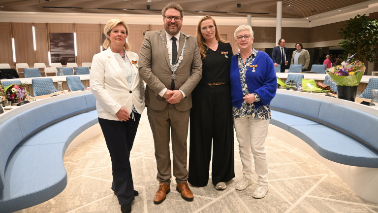 Vier mensen poseren in een moderne vergaderruimte met bloemen op de achtergrond; drie dragen onderscheidingen.