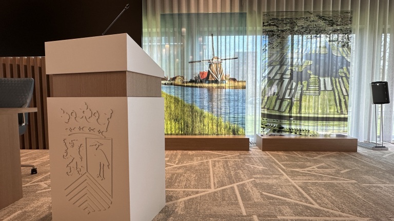 Spreekgestoelte in een vergaderzaal met een afbeelding van een molen en landschap op de achtergrond.