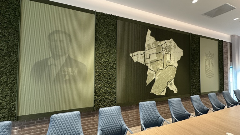 Vergaderruimte met een groene wanddecoratie, waaronder een portret, een plattegrond en een symbool; blauwe stoelen staan bij een houten tafel.