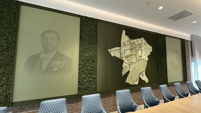 Vergaderruimte met een grote kaart, portret en wapenschild op de muur, en blauwe stoelen rond een houten tafel.