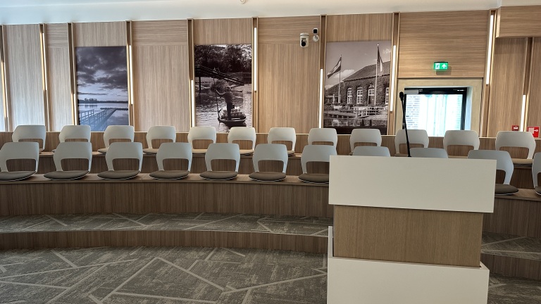 Lege conferentiezaal met houten afwerking, meerdere rijen moderne stoelen, en historische zwart-wit foto's aan de muur.