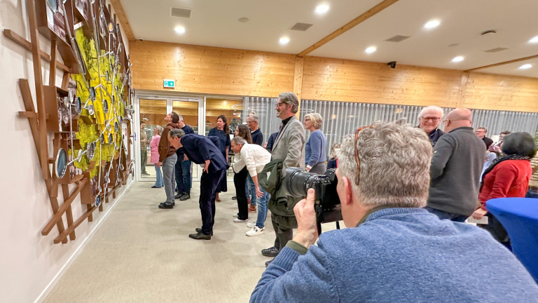 Groep mensen bewondert een grote kunstinstallatie aan de muur in een goed verlichte tentoonstellingsruimte.