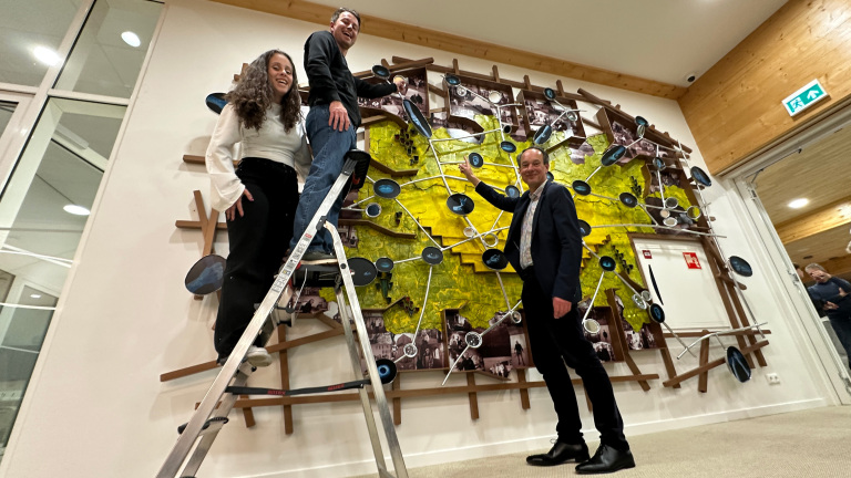 Twee mensen staan op een ladder en een derde persoon staat op de grond voor een kleurrijk kunstwerk aan de muur.