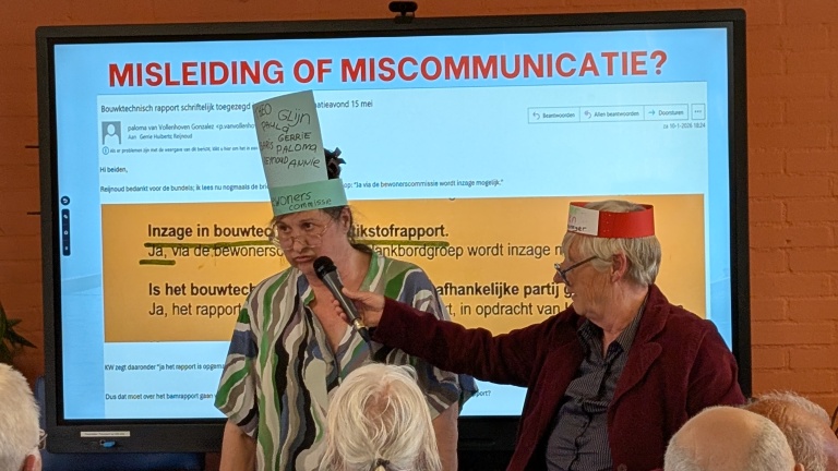 Twee mensen met papieren hoeden staan op een bijeenkomst voor een scherm met de tekst "MISLEIDING OF MISCOMMUNICATIE?".
