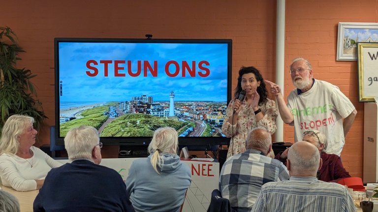 Een vrouw spreekt met een microfoon voor een scherm met "STEUN ONS", terwijl een man wijst; er zitten mensen aan tafel.