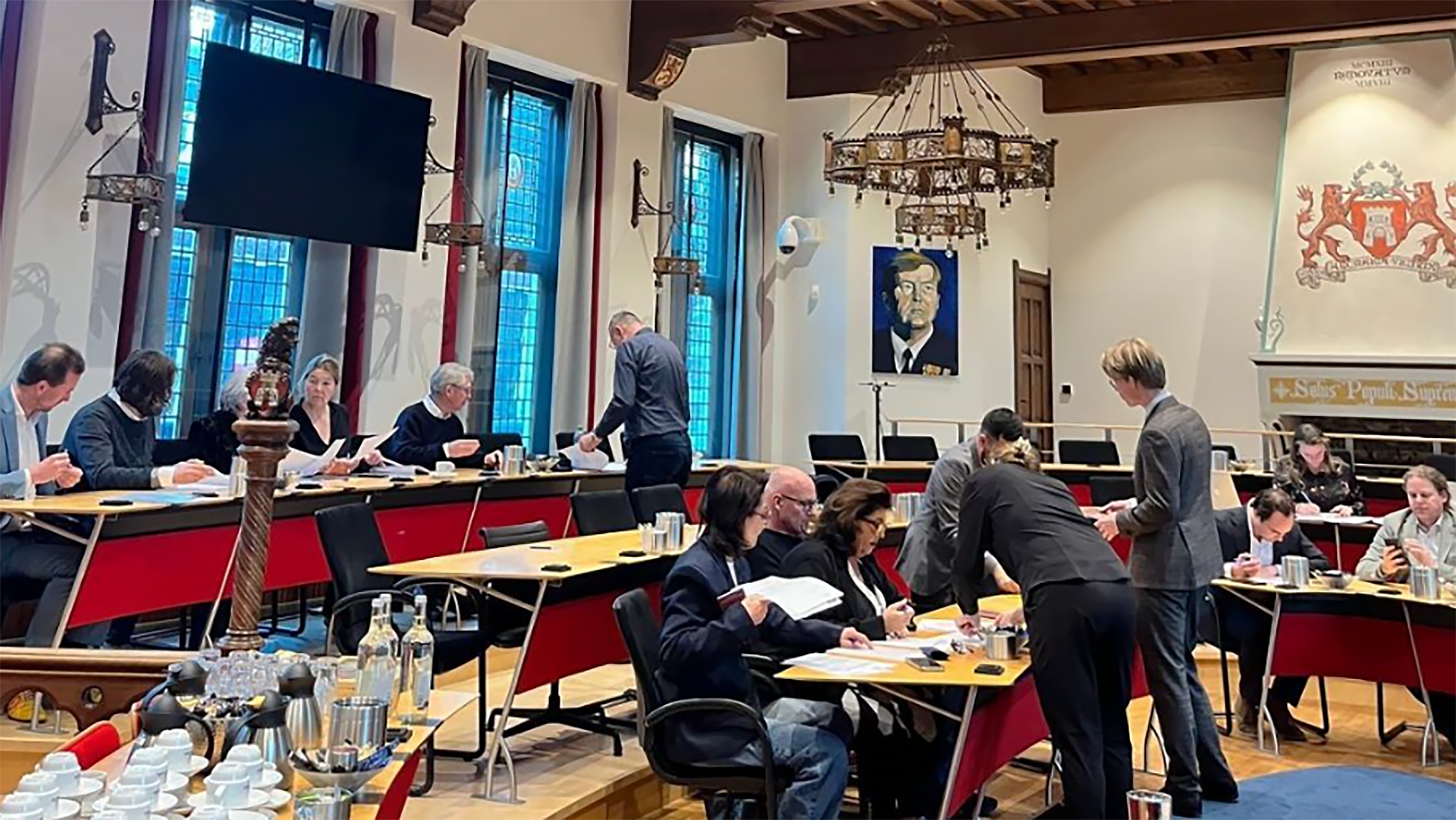 Mensen zitten aan tafels in een vergaderzaal met hoge ramen en een groot schilderij aan de muur.