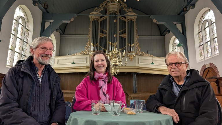 Drie mensen zitten rond een tafel in een kerk met een pijporgel op de achtergrond, glimlachend naar de camera.
