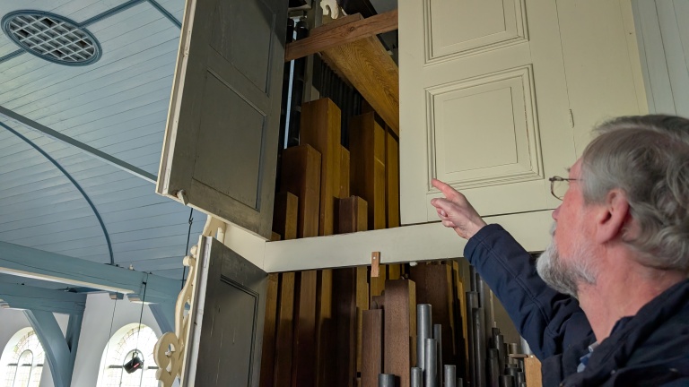 Een man met een bril wijst naar de geopende deuren van een orgelkast met houten orgelpijpen zichtbaar binnenin.