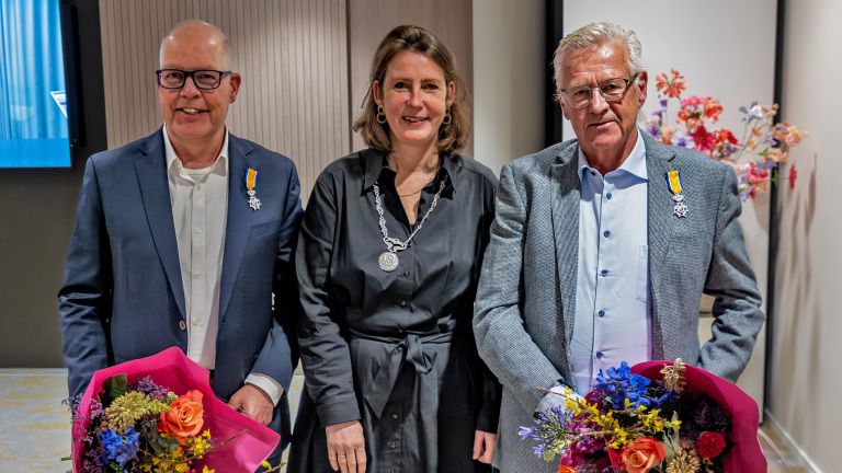 Twee mannen met onderscheidingen en bloemen staan naast een vrouw met een ketting in een binnenruimte.