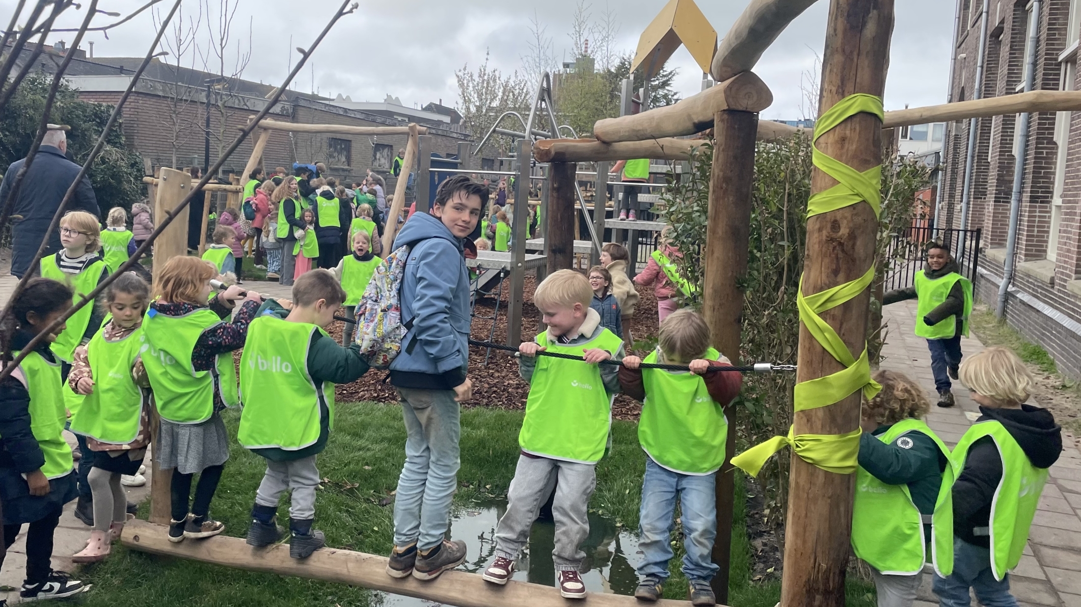 Kinderen in groene hesjes spelen op een houten evenwichtsbalk op een drukke speelplaats.
