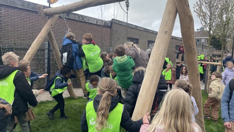 Kinderen spelen in een speeltuin met een grote houten schommel, velen dragen groene hesjes.