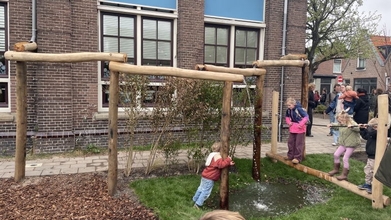 Kinderen spelen op een houten waterspeeltoestel in een tuin bij een bakstenen gebouw.