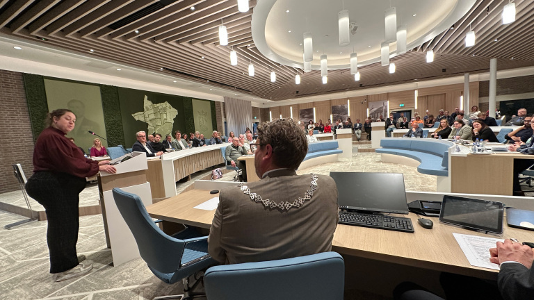 Een vrouw spreekt in een moderne vergaderzaal vol met mensen, met een persoon op de voorgrond die een ambtsketen draagt.