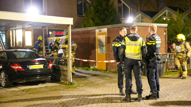 Politieagenten praten buiten een huis terwijl brandweerlieden naar binnen gaan, met een zwarte auto op de oprit.