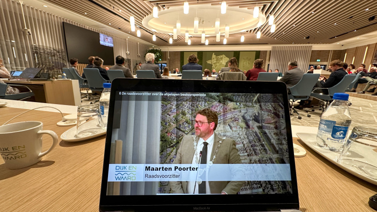 Onder een koepelvormig plafond, een laptop met een afbeelding en tekst over een raadsvoorzitter, omringd door vergadertafels en mensen.